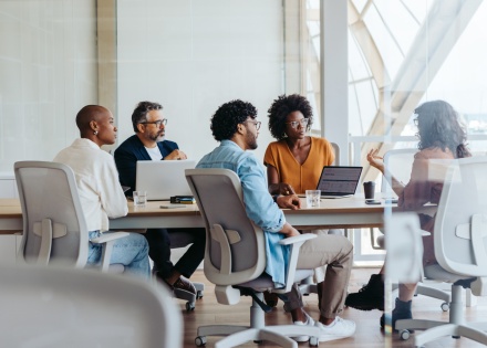 A small diverse workforce collaborates on work tasks while sitting around a conference room table