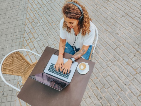 Birds-eye-view of remote worker outdoors, sitting at cafe table with coffee, typing on laptop.