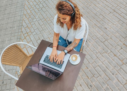 Birds-eye-view of remote worker outdoors, sitting at cafe table with coffee, typing on laptop.