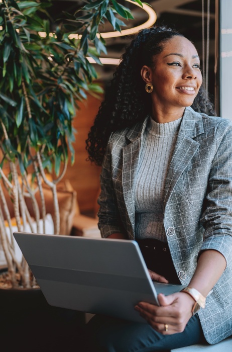 A professional woman smiles while looking out the window while holding a laptop.