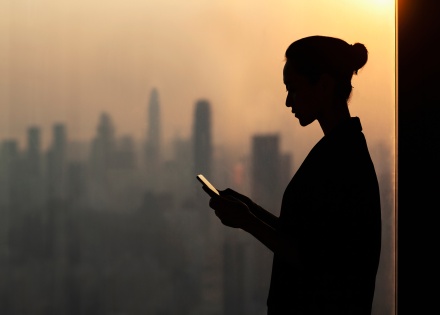Woman working on her mobile phone next to an office window overlooking a city skyline