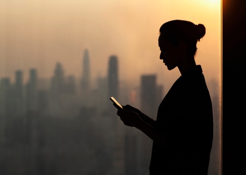 Woman working on her mobile phone next to an office window overlooking a city skyline