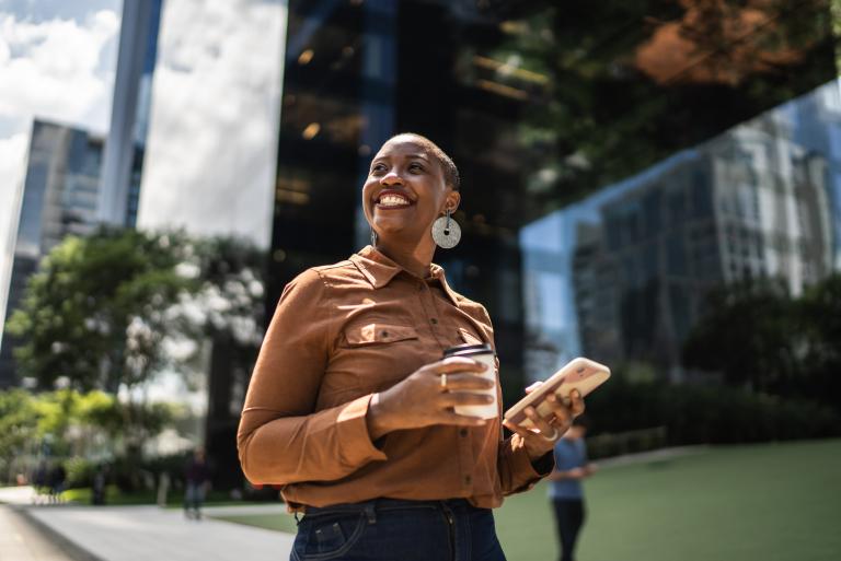 Professional businesswoman holding coffee and phone outside while considering HR compliance.
