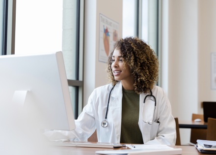 Healthcare provider working on a desktop computer at her desk; wearing a white medical coat and stethoscope
